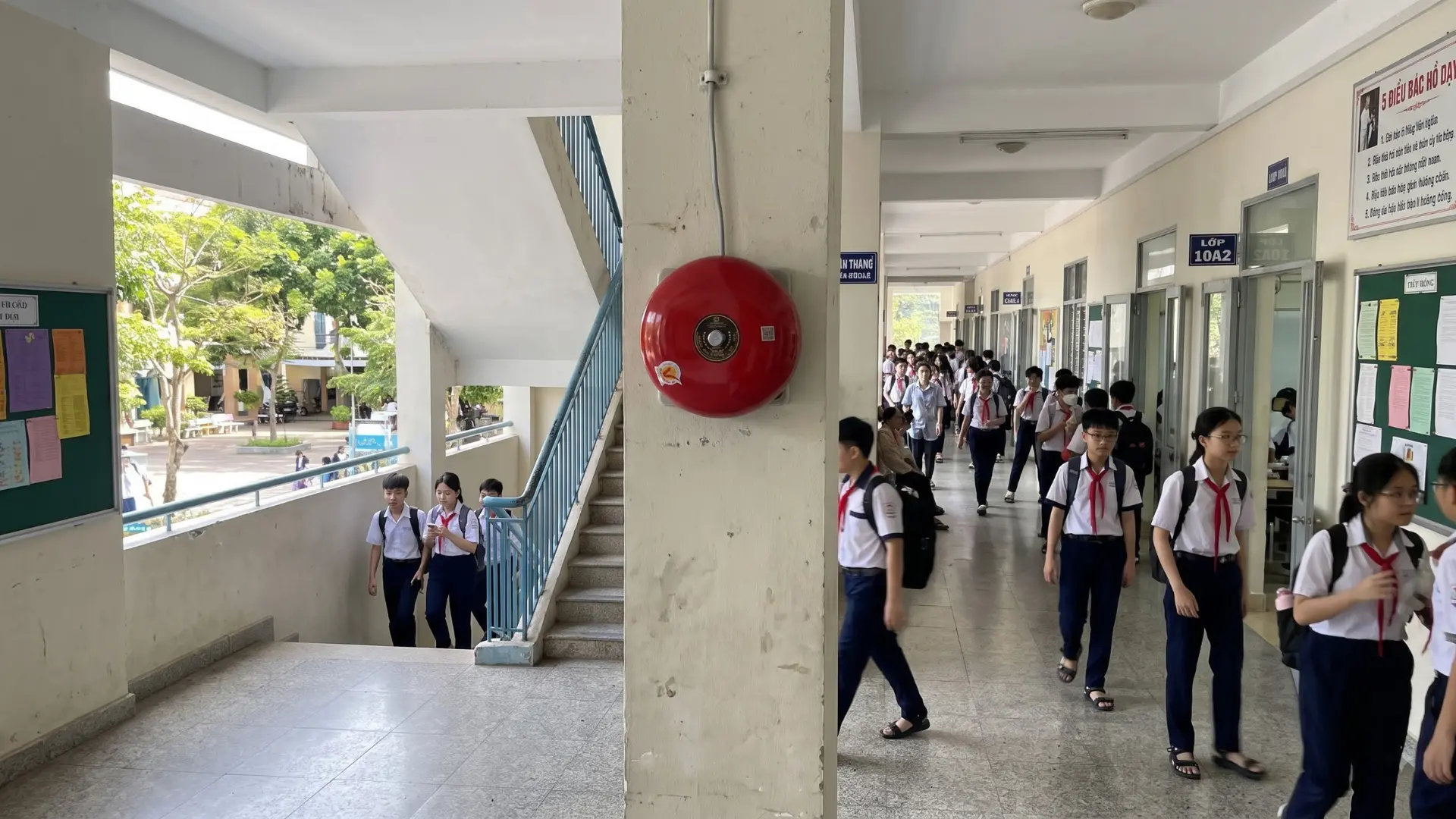 Chuông điện màu đỏ lắp trên cột hành lang trường học, gần cầu thang và dãy phòng học có học sinh di chuyển.
